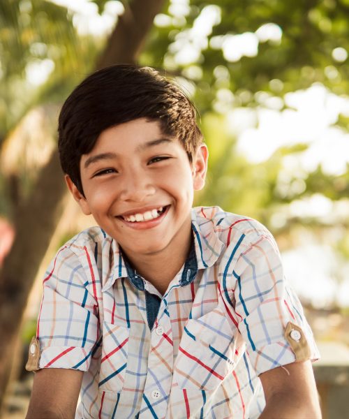 A portrait of a cheerful young boy with a beaming smile, enjoying a sunny day outdoors with a blurred background.